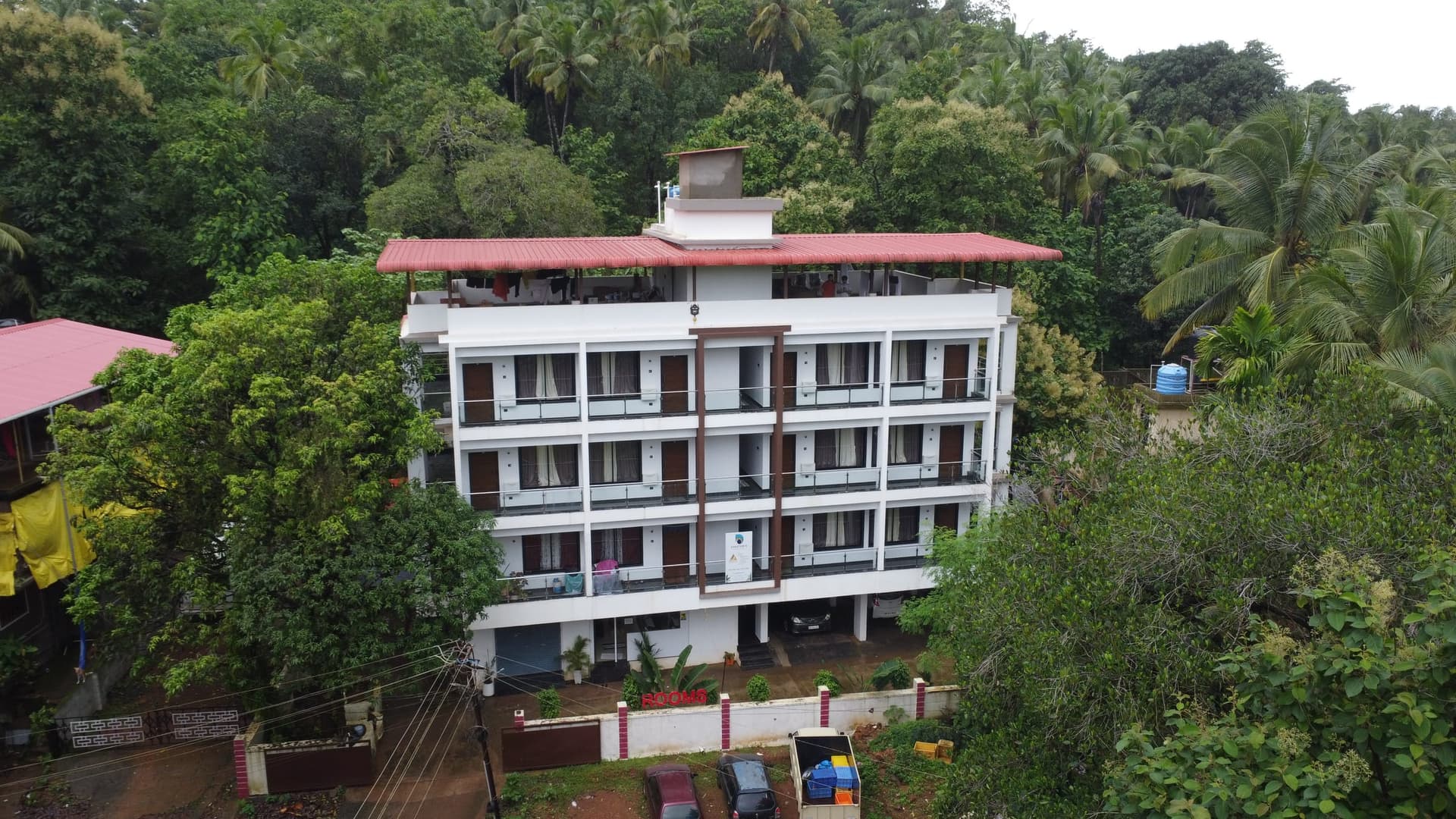 The Palolem Heights house from the air — four storeys of balconies under a red roof, wrapped in palms